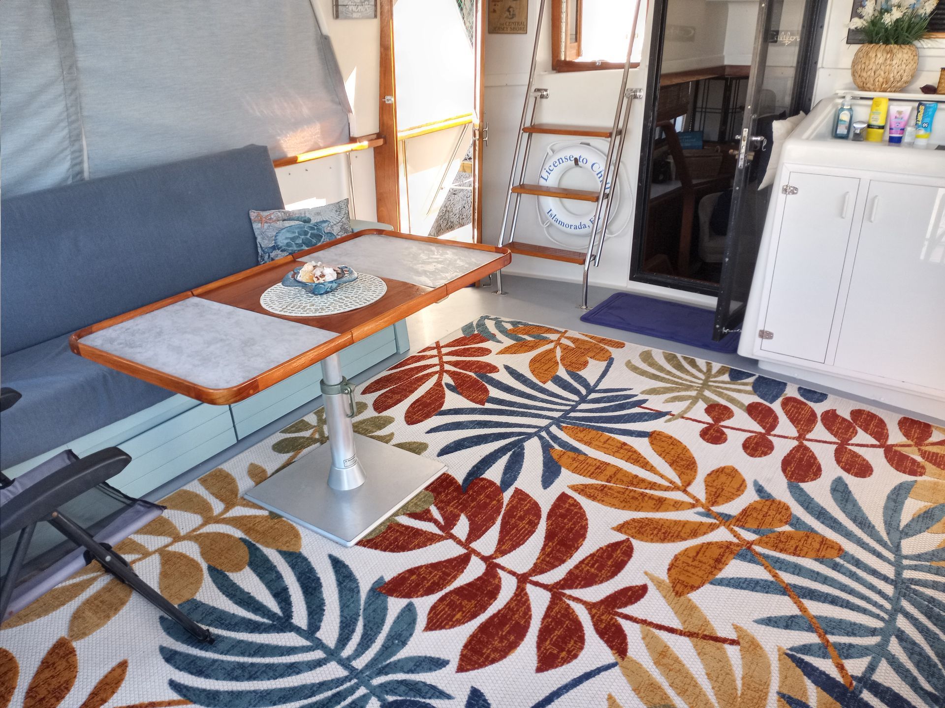 Interior of a boat with a table, sofa, and a colorful leaf patterned rug.