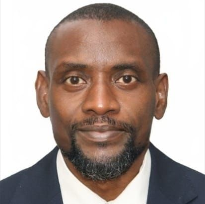 Man in a suit, dark complexion, looking at the viewer. He has a short beard and a red tie.