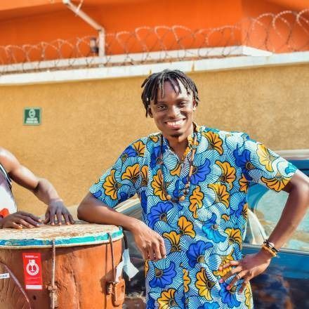 Young man in blue patterned shirt, smiling, leaning on a drum.