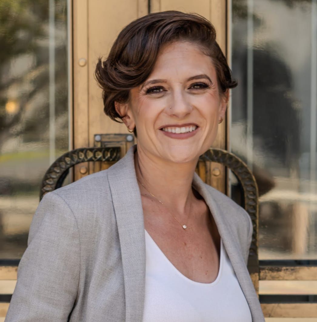 Woman with short brown hair, smiling, in front of a gold door, wearing a blazer and white shirt.