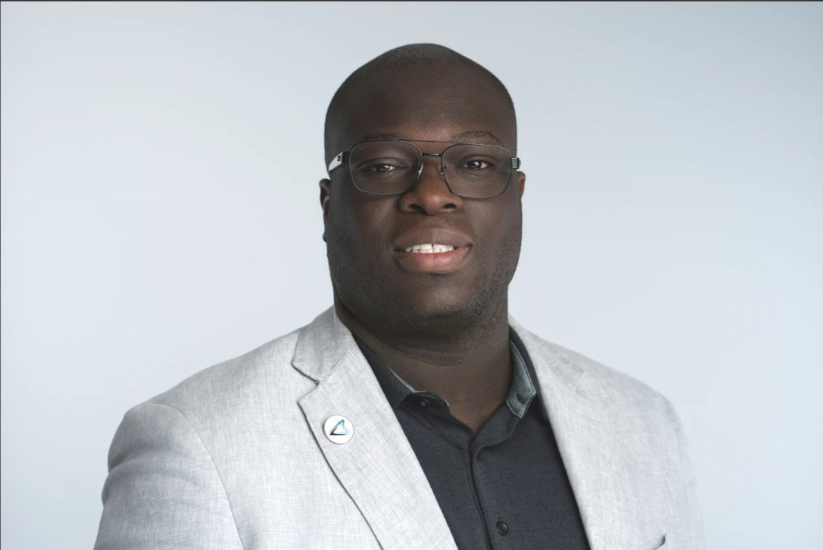 Man with glasses wearing a light gray blazer and black shirt, smiling at the camera. White background.