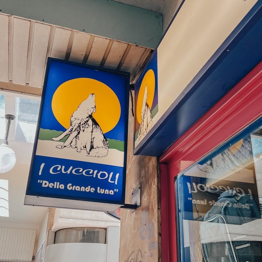 Insegna blu sopra una porta, con il disegno di un sole giallo e di una montagna e la scritta «I cuccioli».