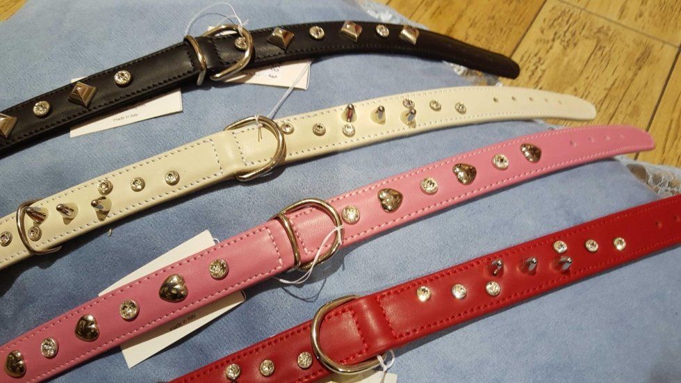 Quattro cinture di pelle borchiate nere, bianche, rosa e rosse, disposte in diagonale su un tessuto.