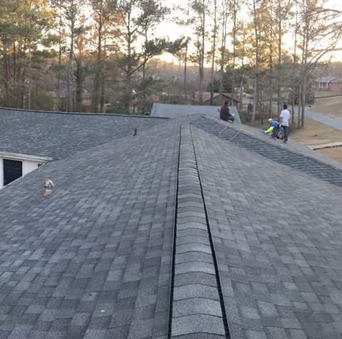 Workers on a gray shingle roof, preparing materials against a backdrop of trees and a house.