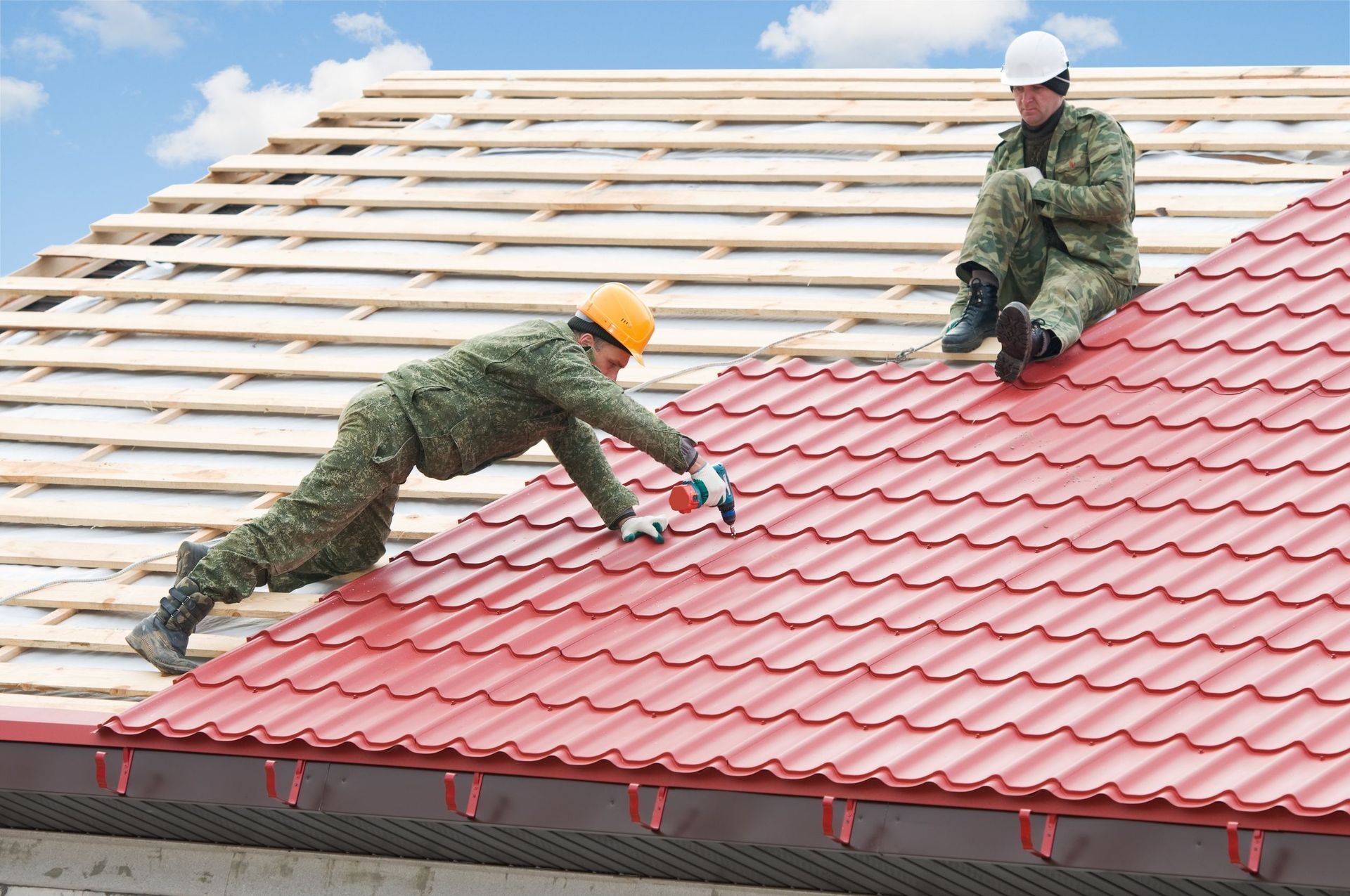 Two roofers in camouflage outfits installing red metal roofing on a house under a blue sky.