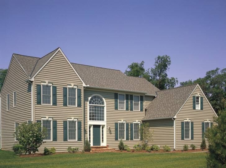 Two-story house with tan siding, green shutters and trim, and a blue sky.