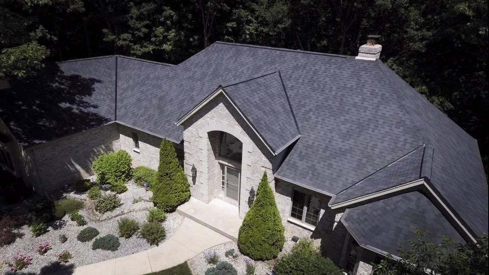 Stone house with dark gray shingle roof, surrounded by trees and landscaping.