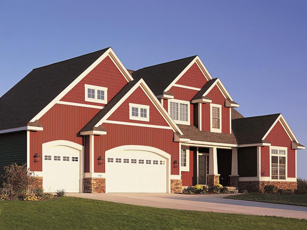 Red sided house with white garage doors, lawn, and blue sky.
