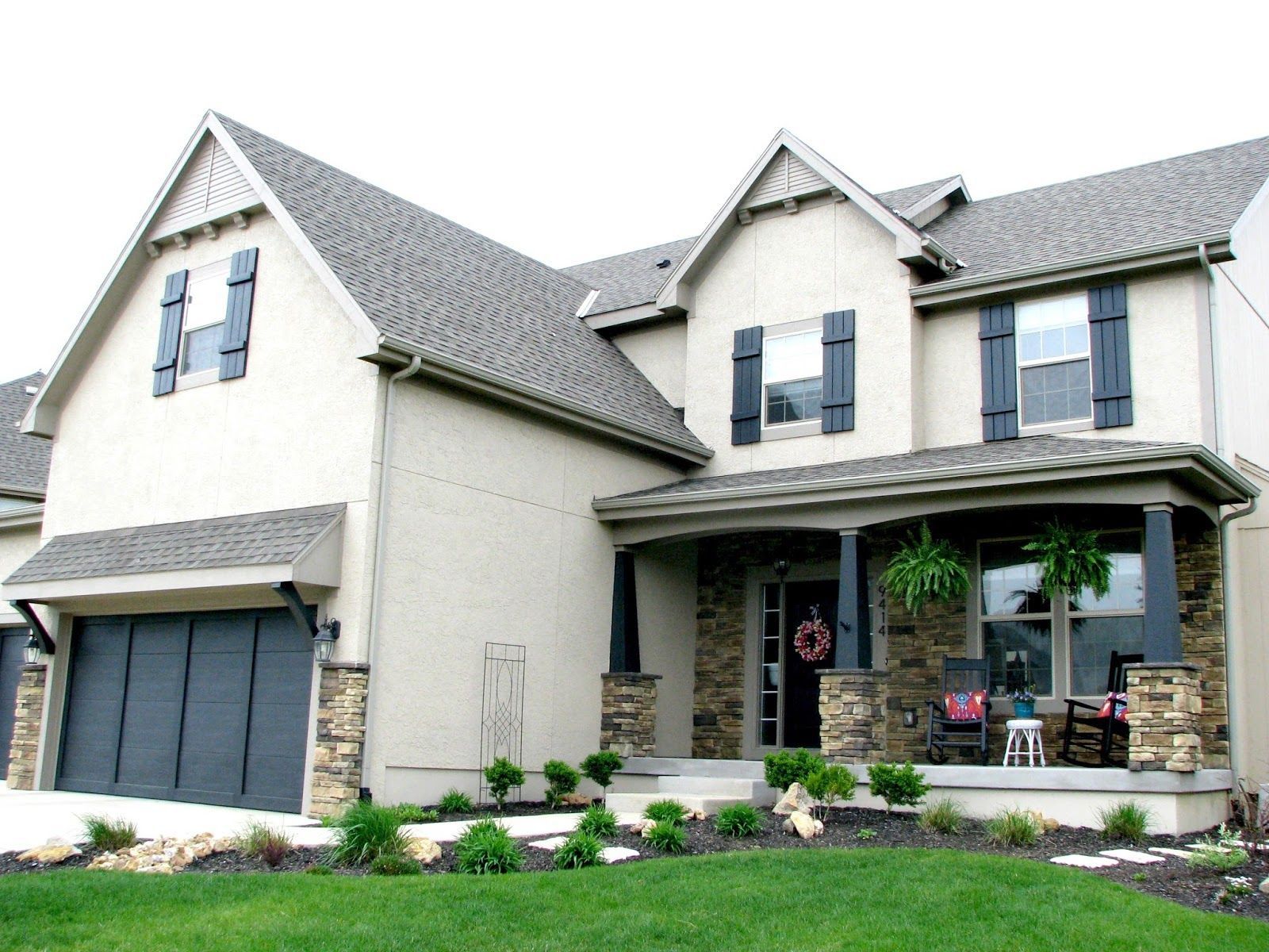 Two-story house with tan stucco exterior, gray roof, dark shutters, and stone accents.