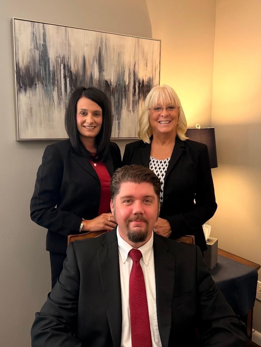 Three colleagues posing in a warmly lit office, two women standing behind a seated man in a black suit and red tie.
