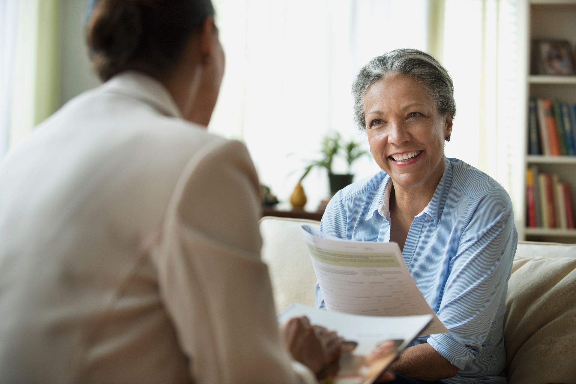 A woman is sitting on a couch talking to another woman while holding a piece of paper.