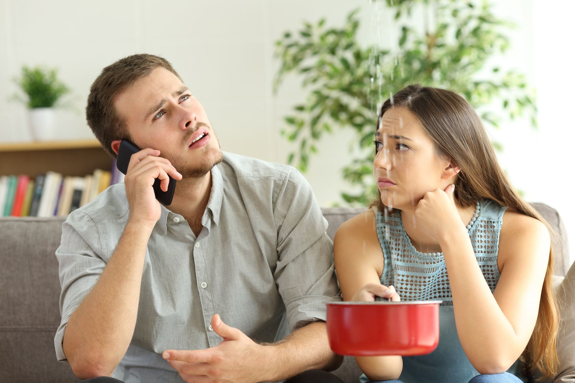 A man is talking on a cell phone while a woman looks at a pot of water coming out of the ceiling.