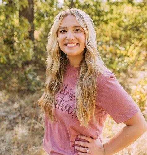 Smiling woman in a pink shirt standing outdoors with hand on hip in a sunlit wooded area