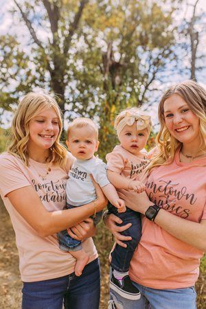 Two women holding toddlers outdoors in matching peach shirts, smiling in front of trees