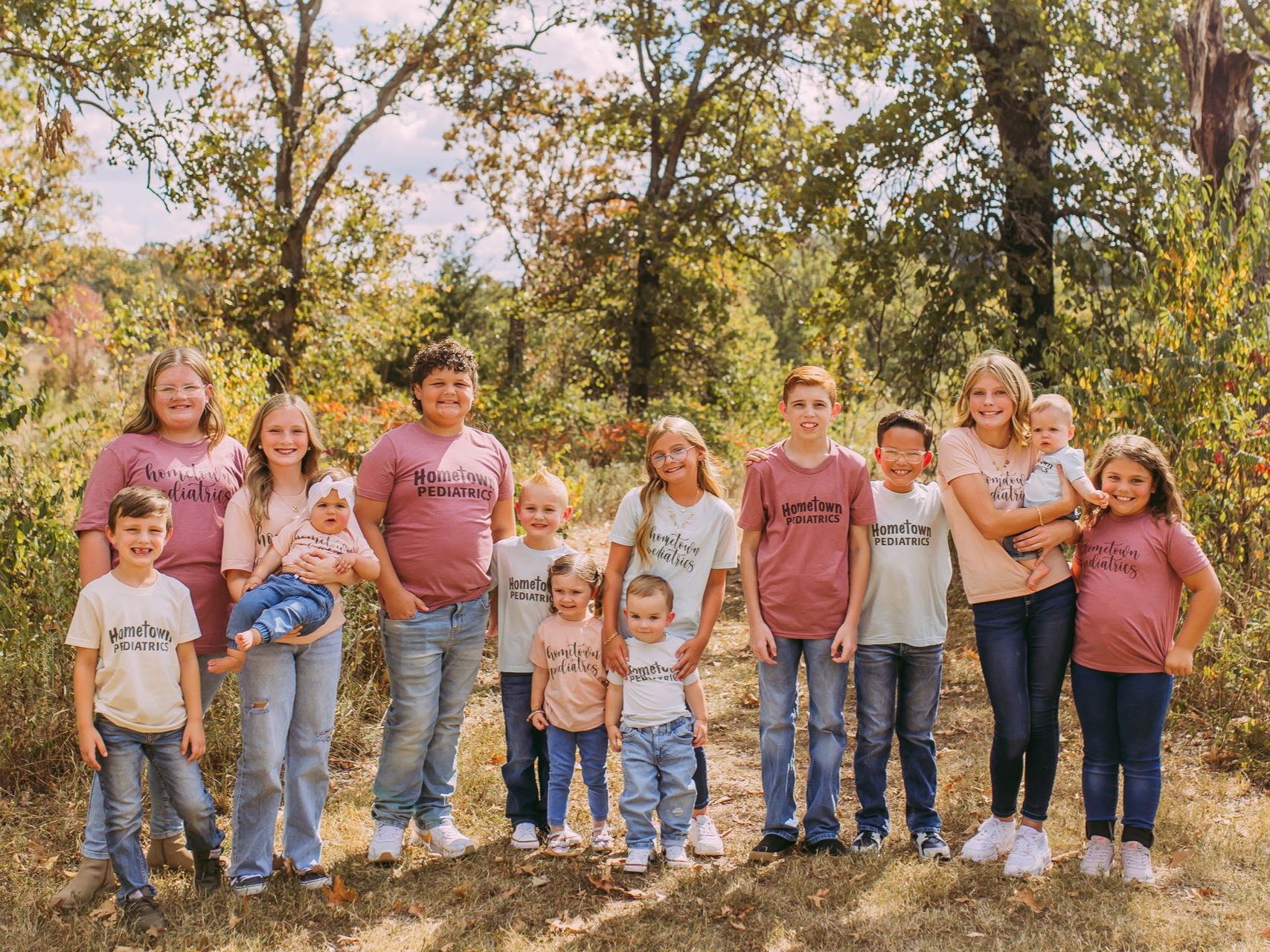 Group of people standing outdoors among trees, posing together in a sunny woodland setting.