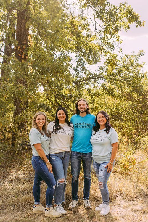 Four friends standing together outdoors in front of trees, smiling at the camera.