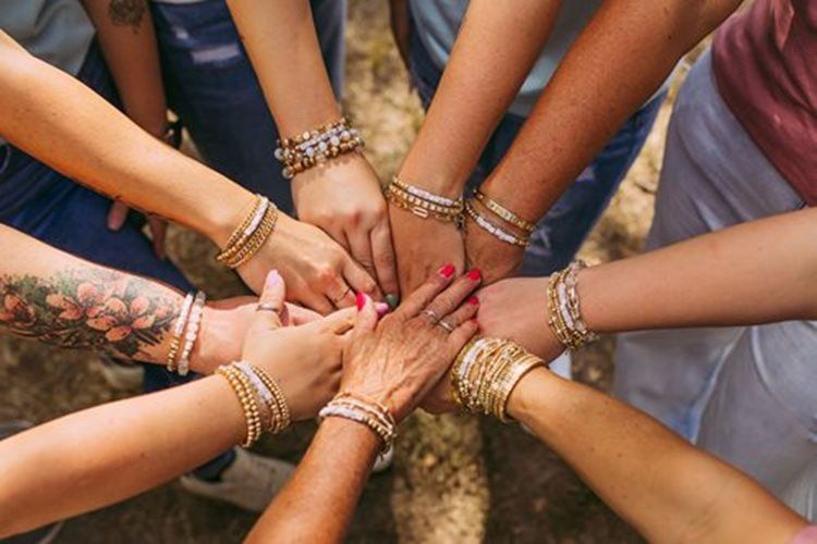 Multiple hands with bracelets stacked together in a circle on a wooden surface