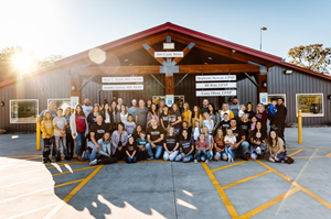 Large group posing outside a red-roofed building in a sunlit parking lot