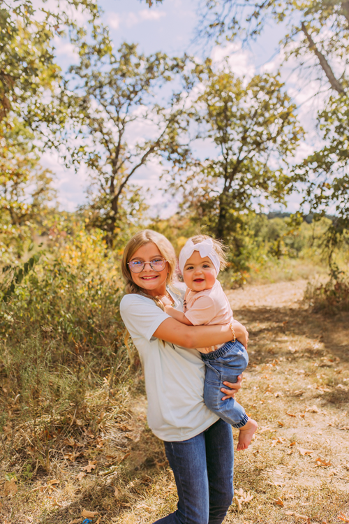 Person holding a smiling child outdoors on a sunny path with trees and grass
