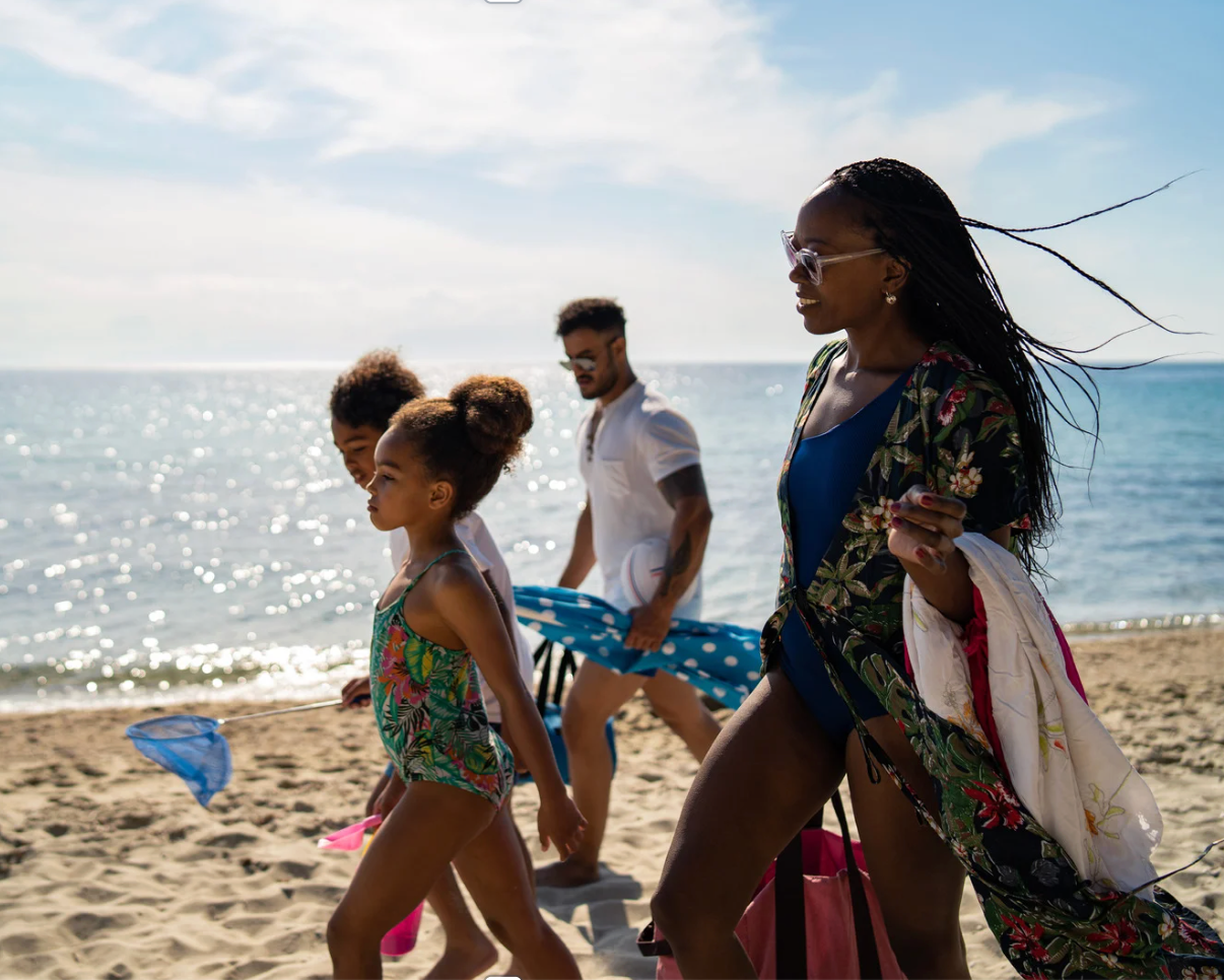Family walking along a sunny beach beside the ocean, with colorful flags in the sand