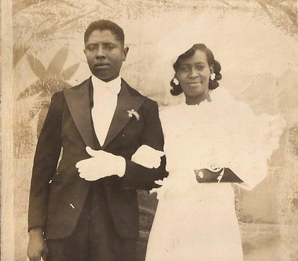 Studio portrait of a formally dressed Aunt Alicia's Inn and her husband Bobo standing arm-in-arm, sepia-toned background.