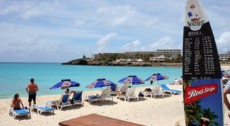 Sunny beach with blue umbrellas, lounge chairs, turquoise water, and a resort menu board on the right.