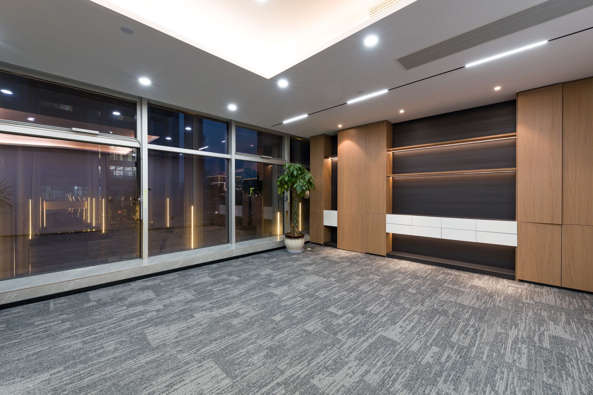 Empty modern office lobby with large windows, gray carpet, wood paneling, and recessed ceiling lights