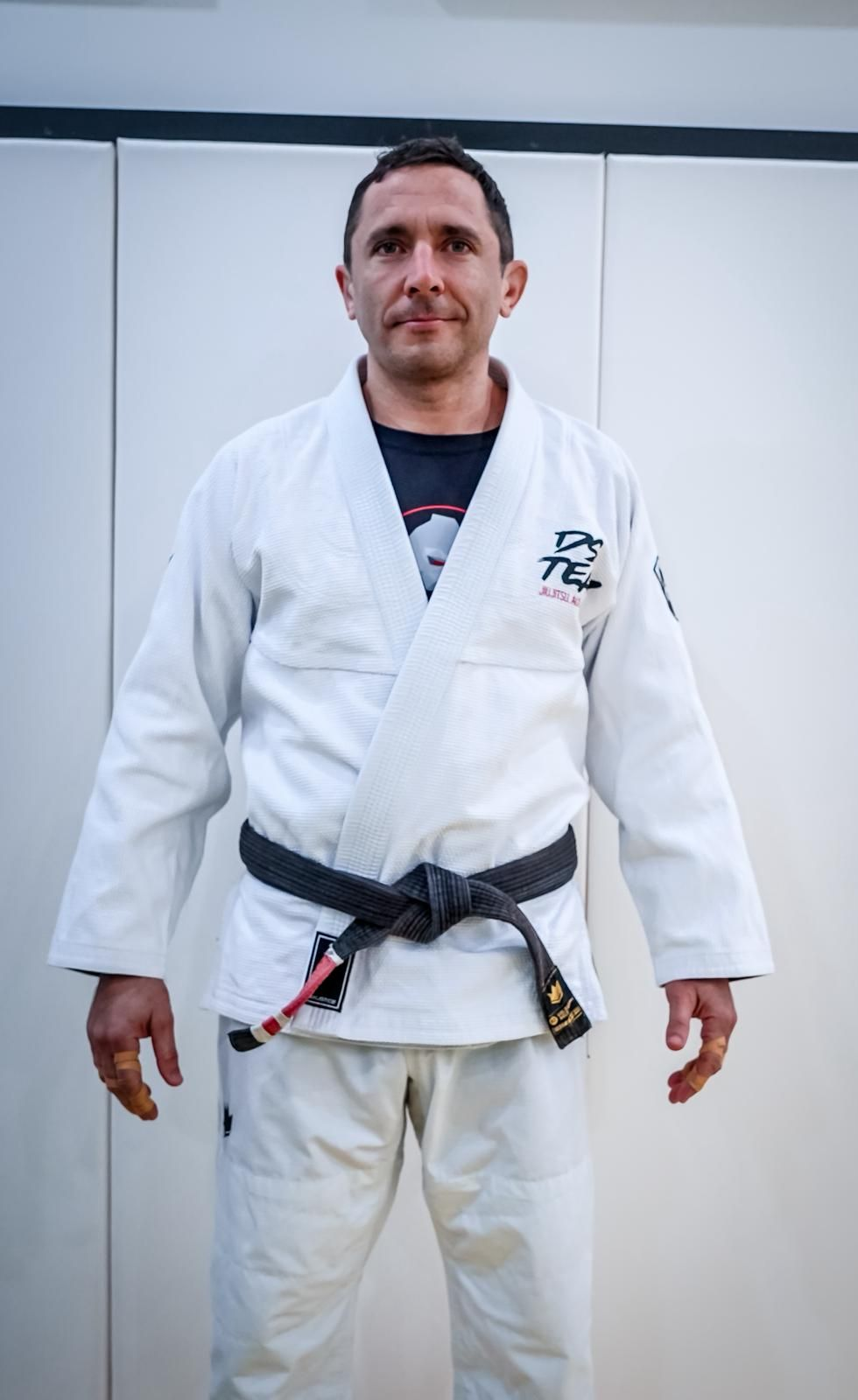 Man in a white gi with black belt standing indoors against a plain wall.