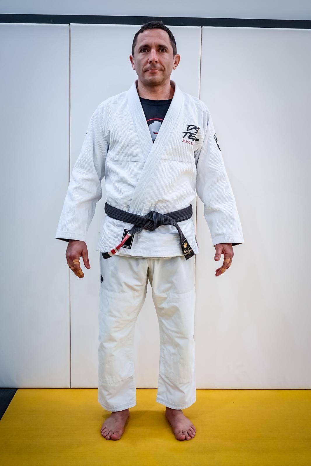 Person in white martial arts gi and black belt standing on a yellow mat in front of white wall panels.