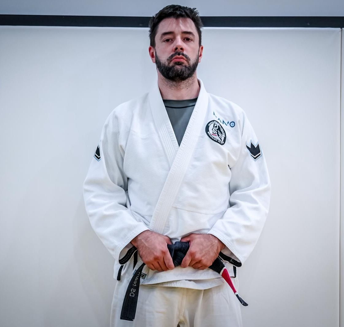 Man in white martial arts gi with black belt standing indoors against a plain wall