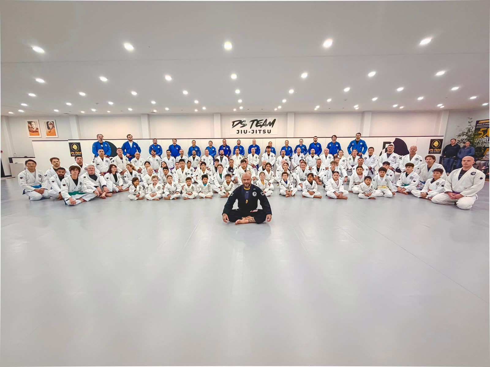 Martial arts group photo with students in white and blue uniforms posing in a dojo, teacher seated front center