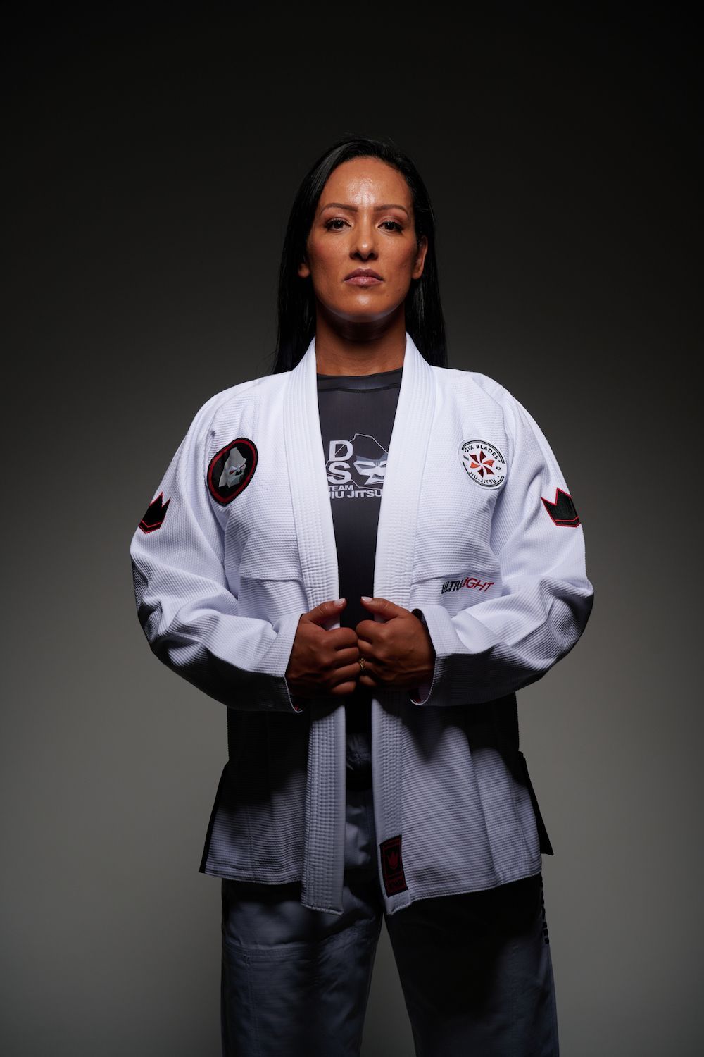 Woman in a white martial arts gi stands against a black background, hands clasped at her waist.