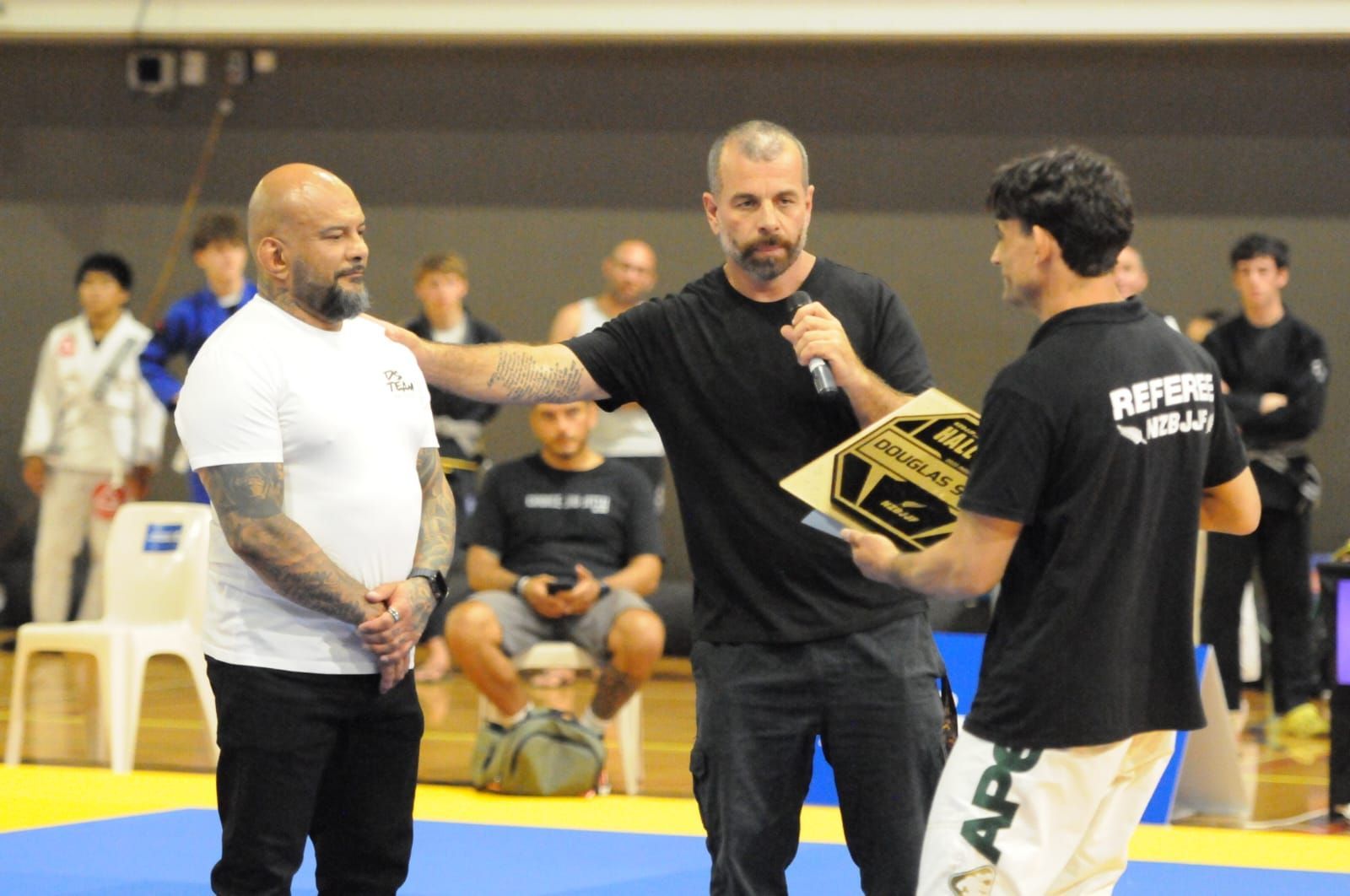 Two men face a referee in a martial arts gym, with spectators seated behind them.