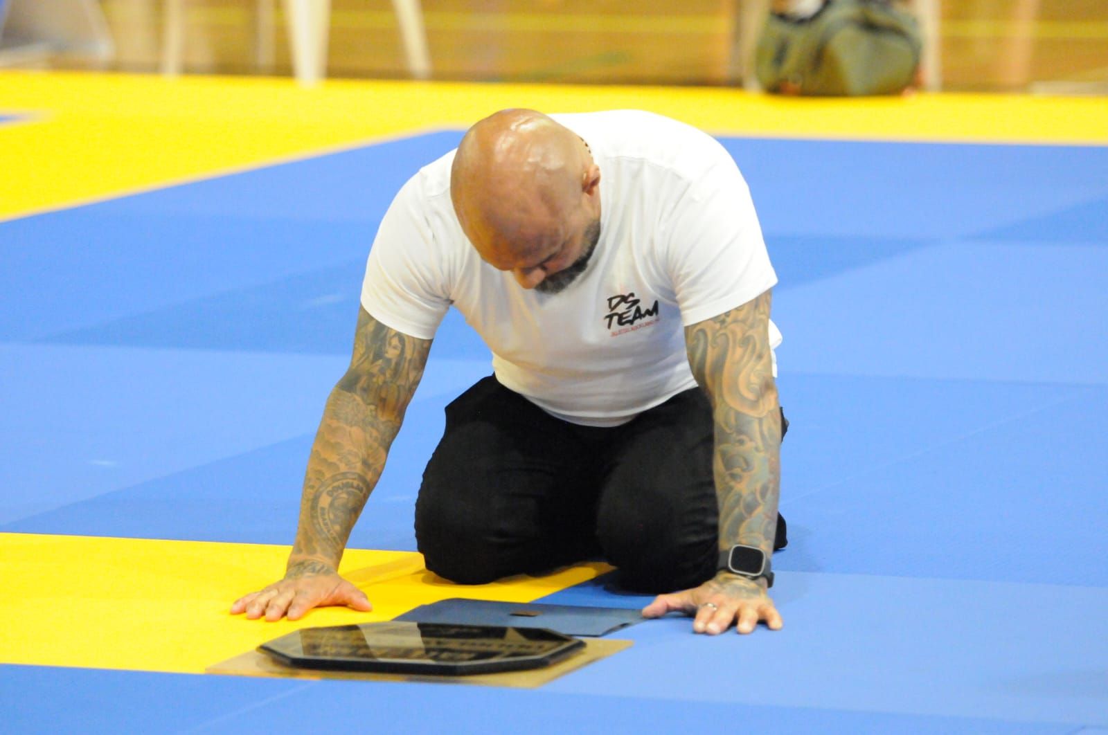 Man in white shirt kneels on blue mat beside a tablet in a gym or dojo
