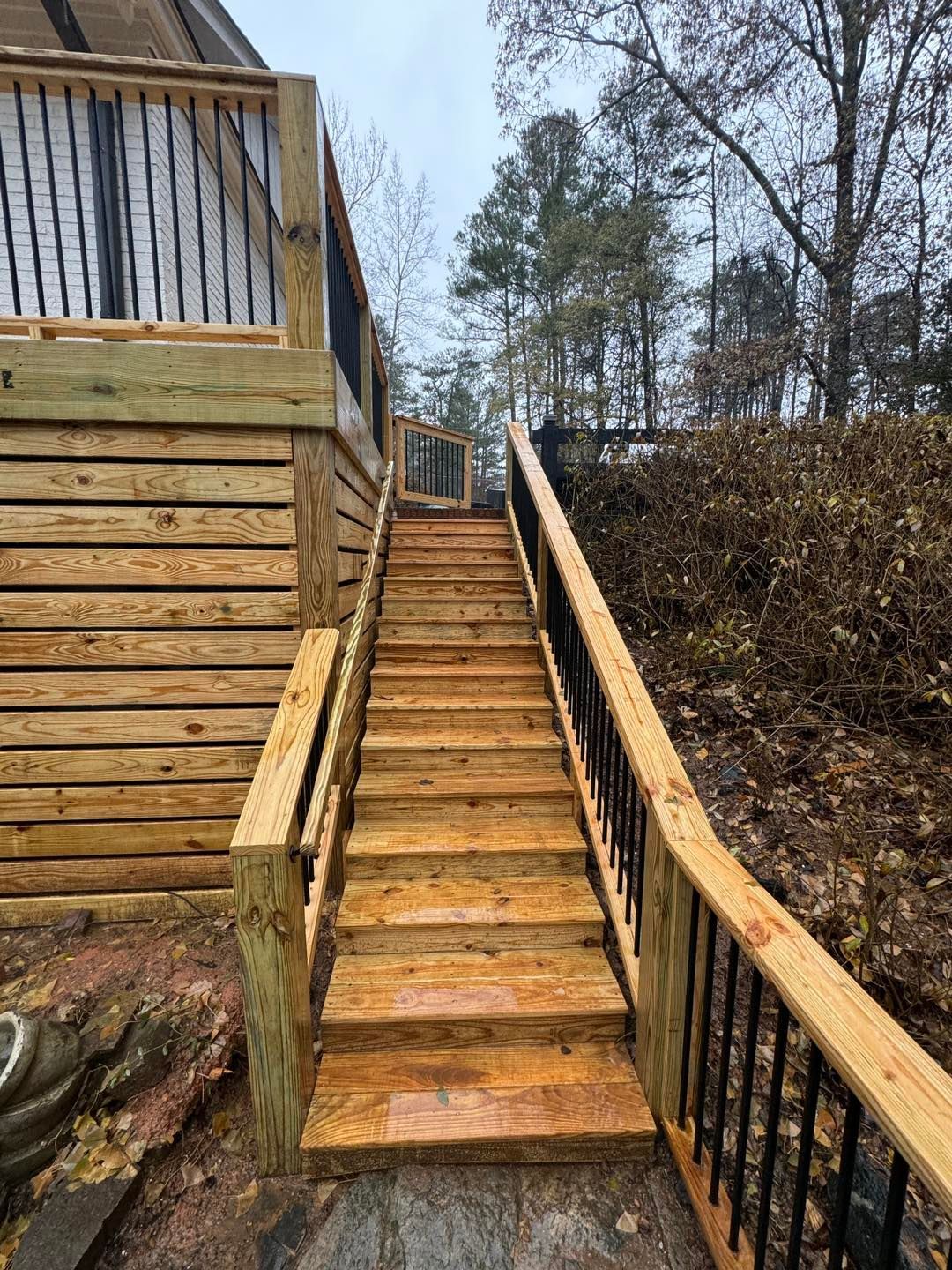 A wooden staircase with black metal railings ascends to an elevated deck next to a wooded hillside.