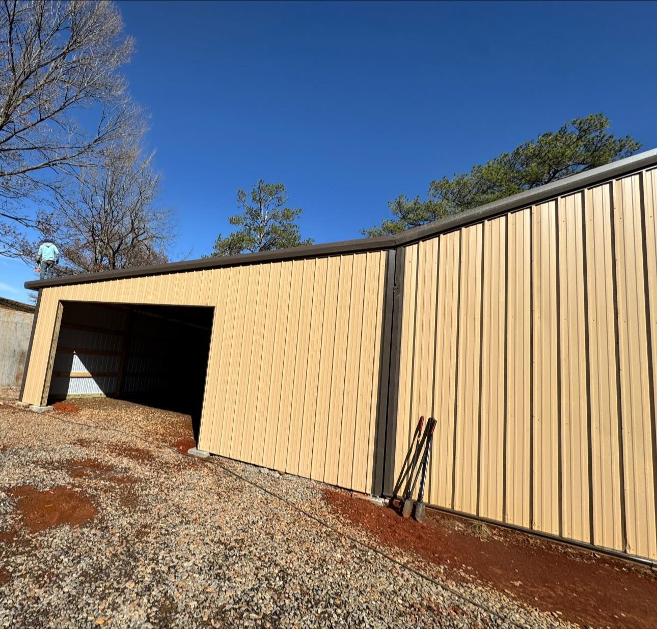 A tan, metal-sided building with an open garage bay set on a gravel lot under a clear blue sky.