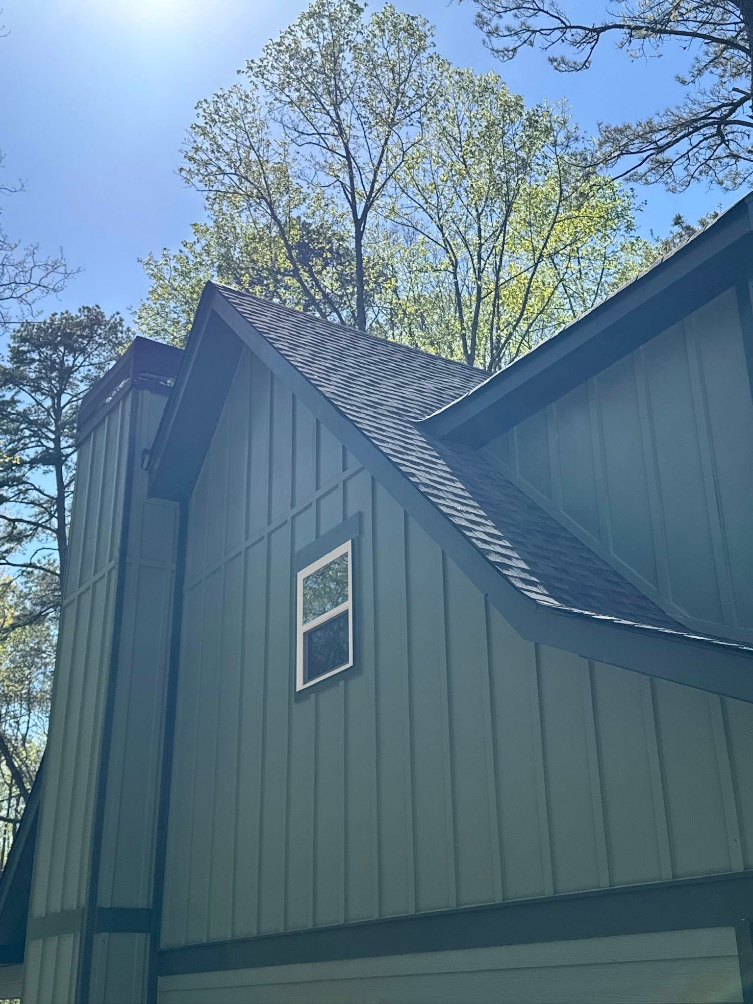 Low-angle view of a sage-green house with vertical wood siding, a square window, and a dark shingled roof under a sunny sky.