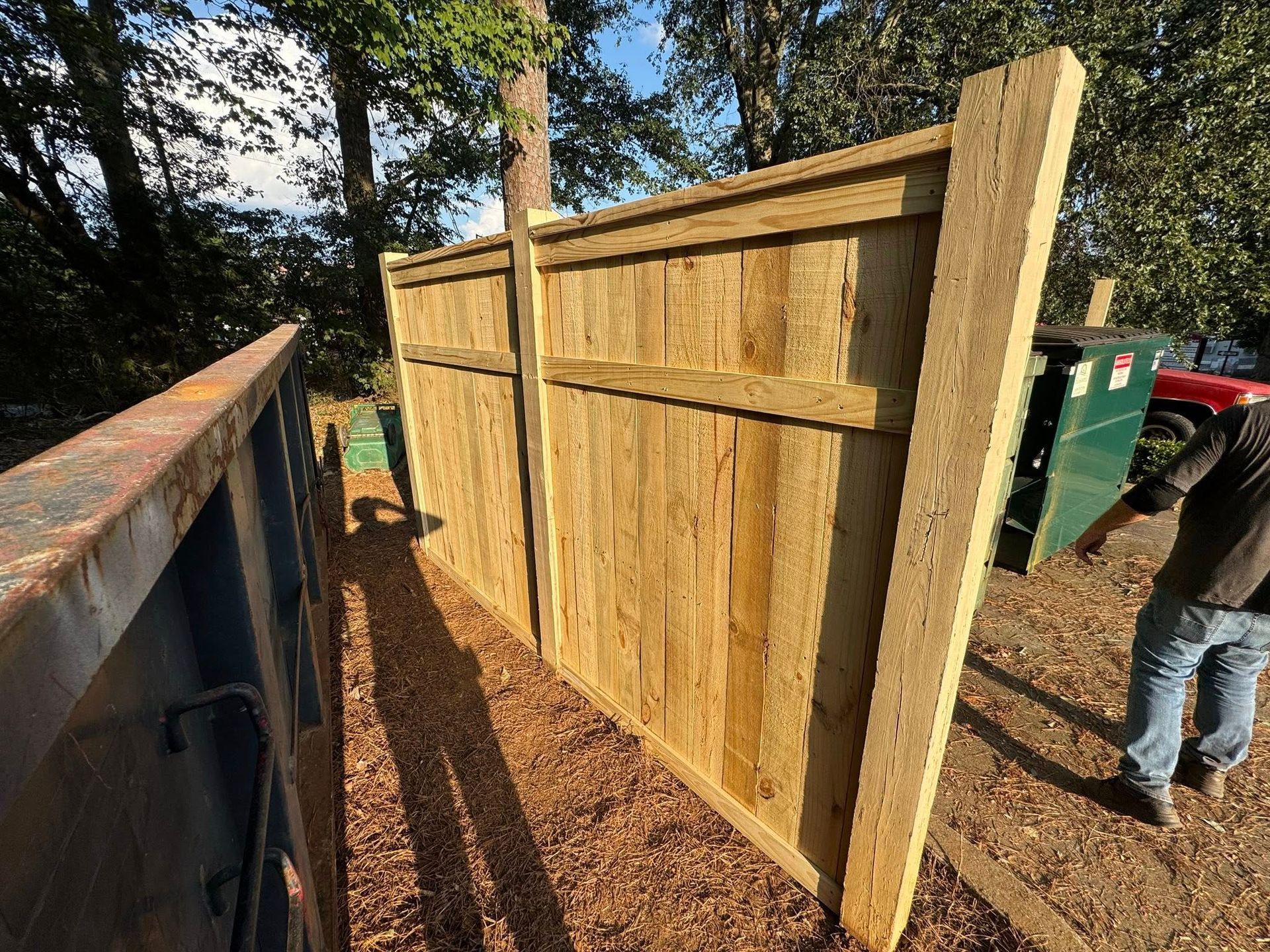A tall wooden fence stands in an outdoor area with wood chips on the ground near a dumpster.