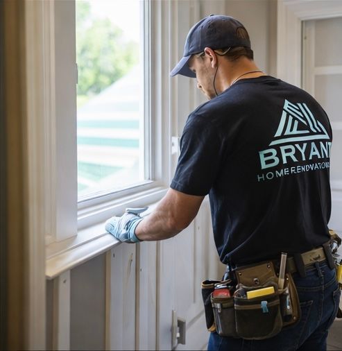 A person in a Bryant Home Renovations shirt and tool belt works on installing window trim.
