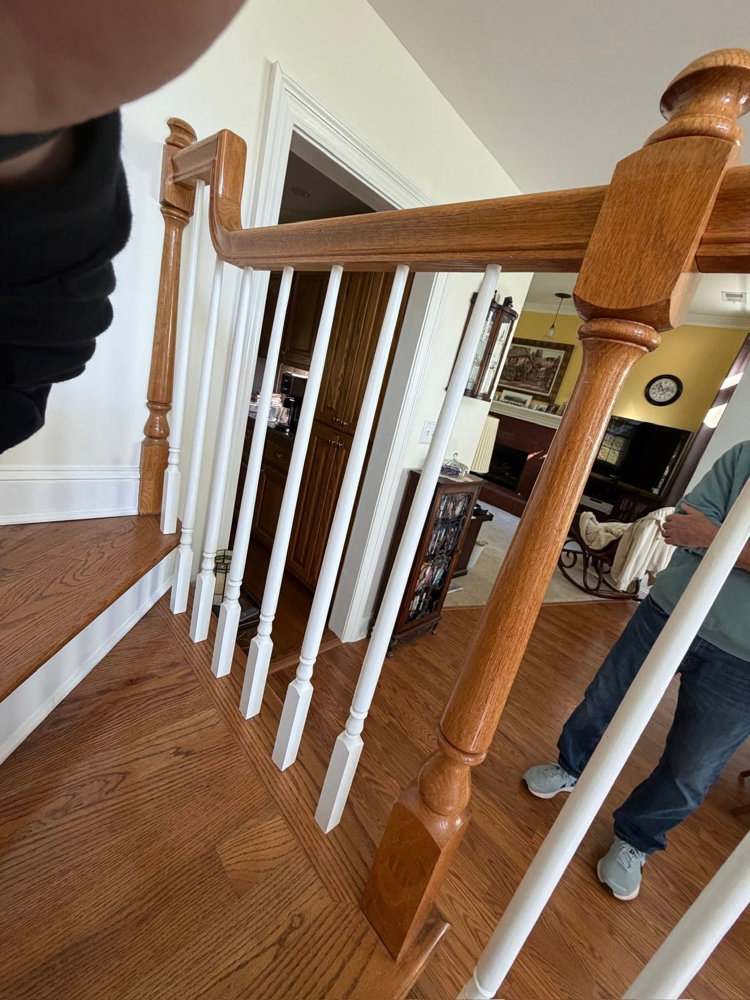 A wooden staircase railing with white balusters in an indoor setting, with a person standing partially visible nearby.