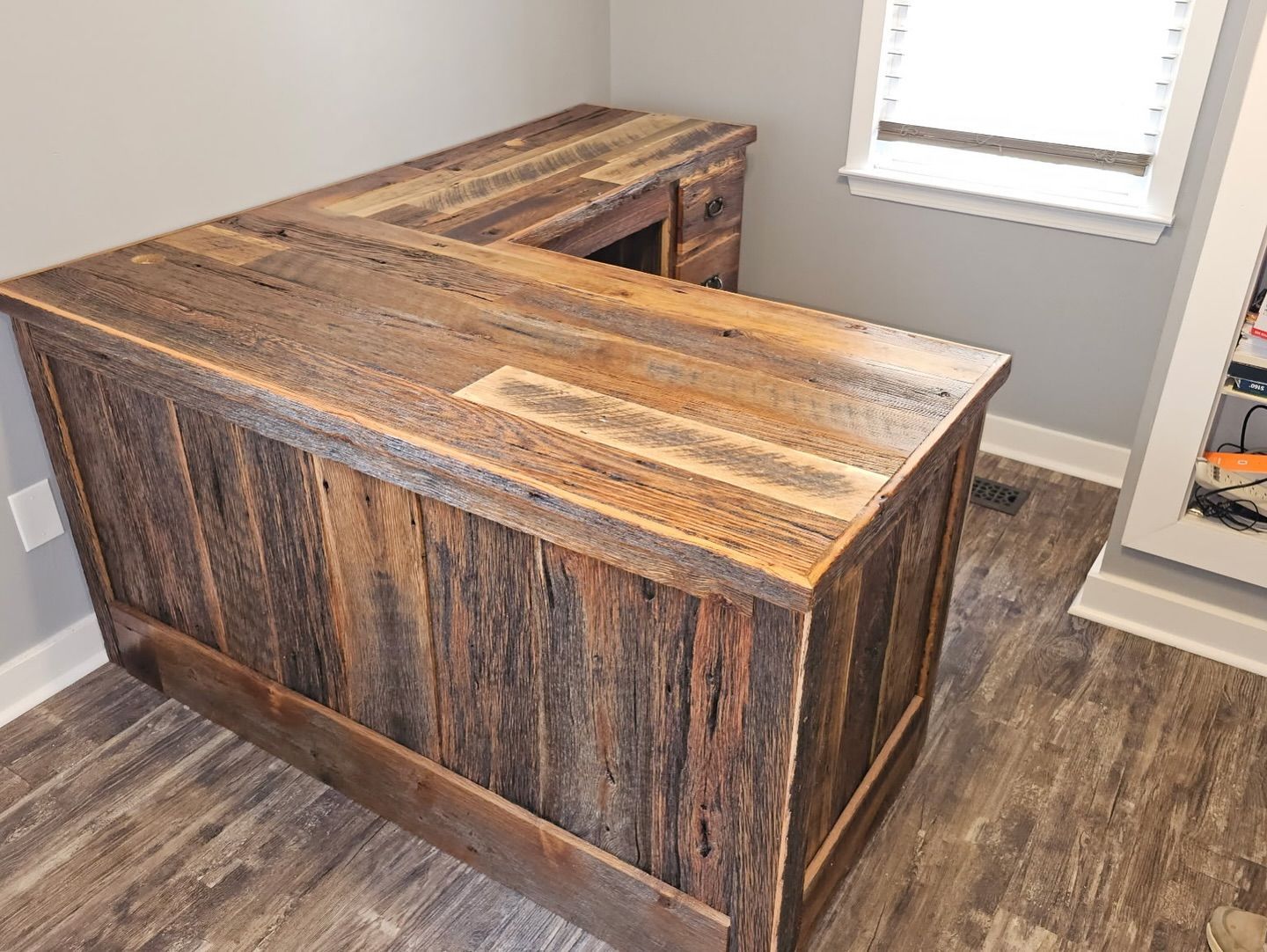 L-shaped rustic wooden desk made of reclaimed timber in a home office with gray walls and wood-patterned flooring.