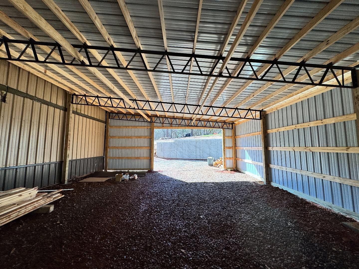 Interior view of a pole barn under construction with gravel floors, metal siding, and open wood-truss ceiling beams.