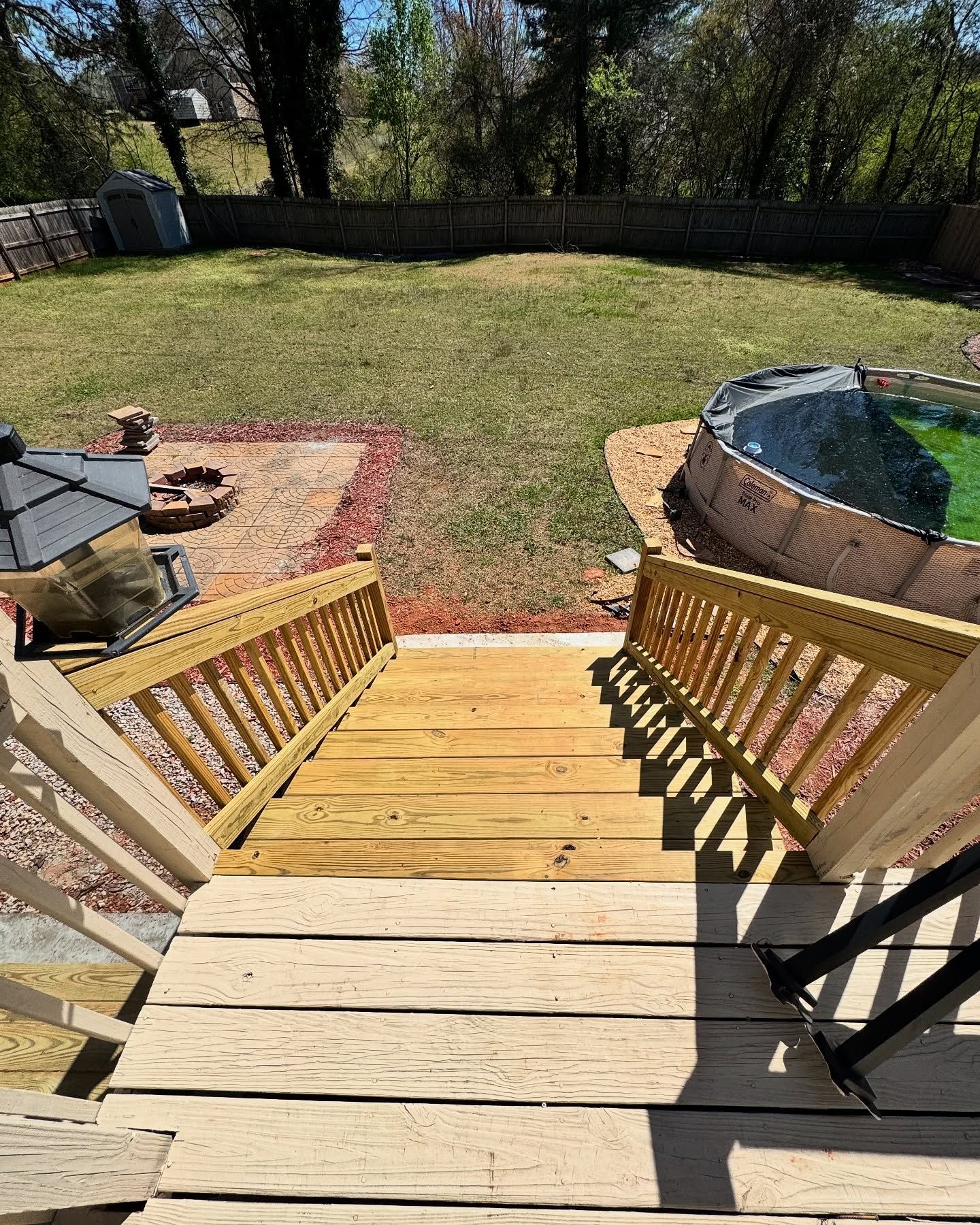 A wooden deck and staircase looking down into a sunny backyard with a pool and a circular fire pit area.