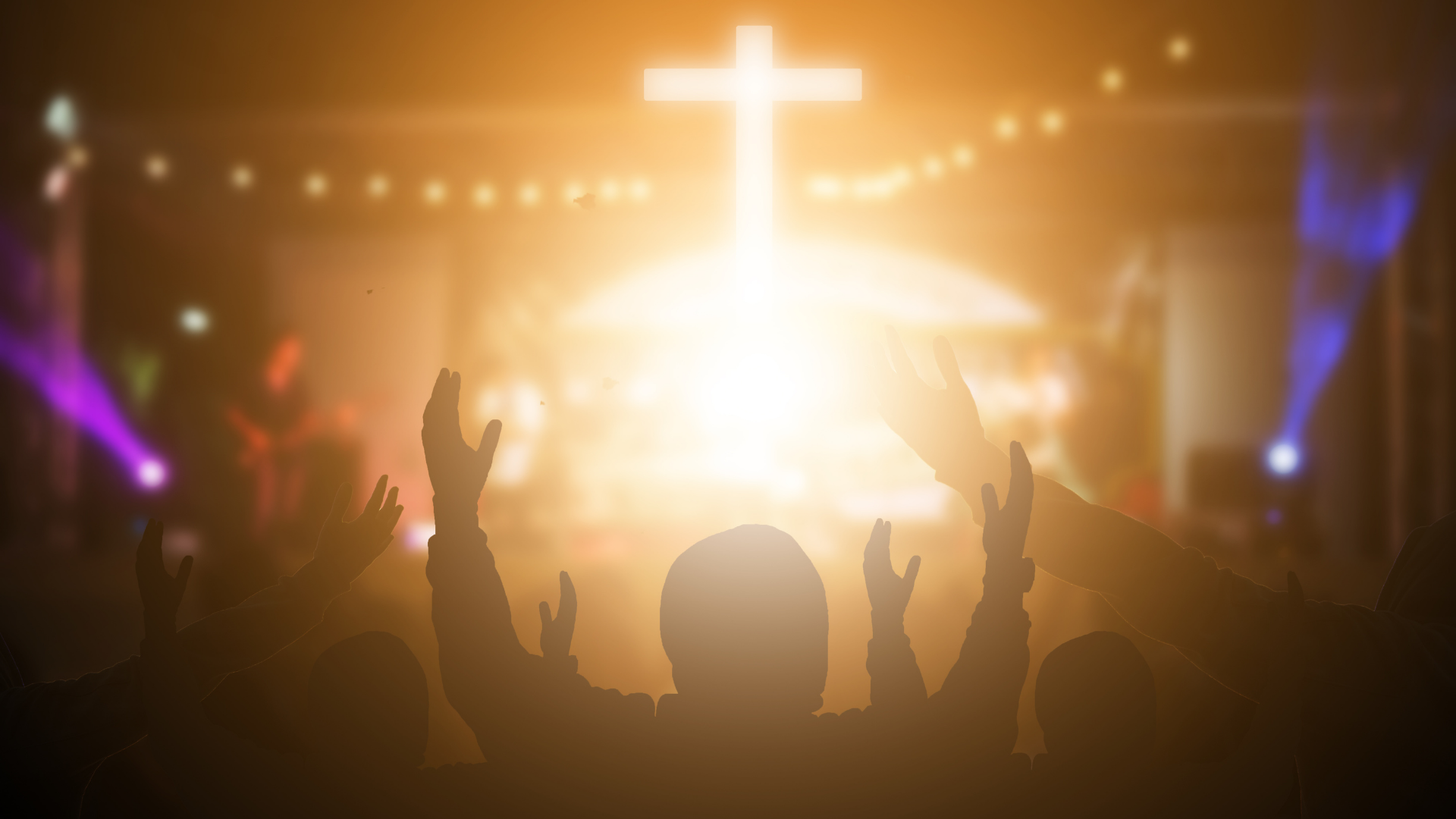 Crowd with raised hands facing a glowing cross on stage under purple and gold lights