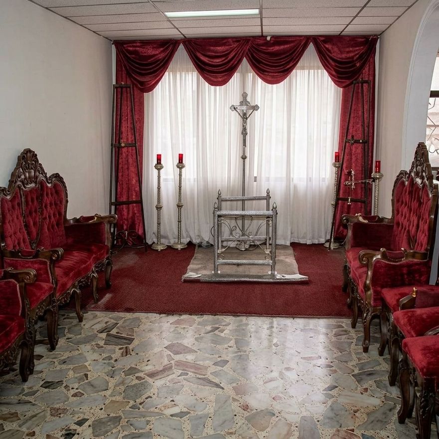 Pequeña capilla con sillas de terciopelo rojo, cortinas blancas, altar y crucifijo bajo cortinas burdeos ornamentadas.