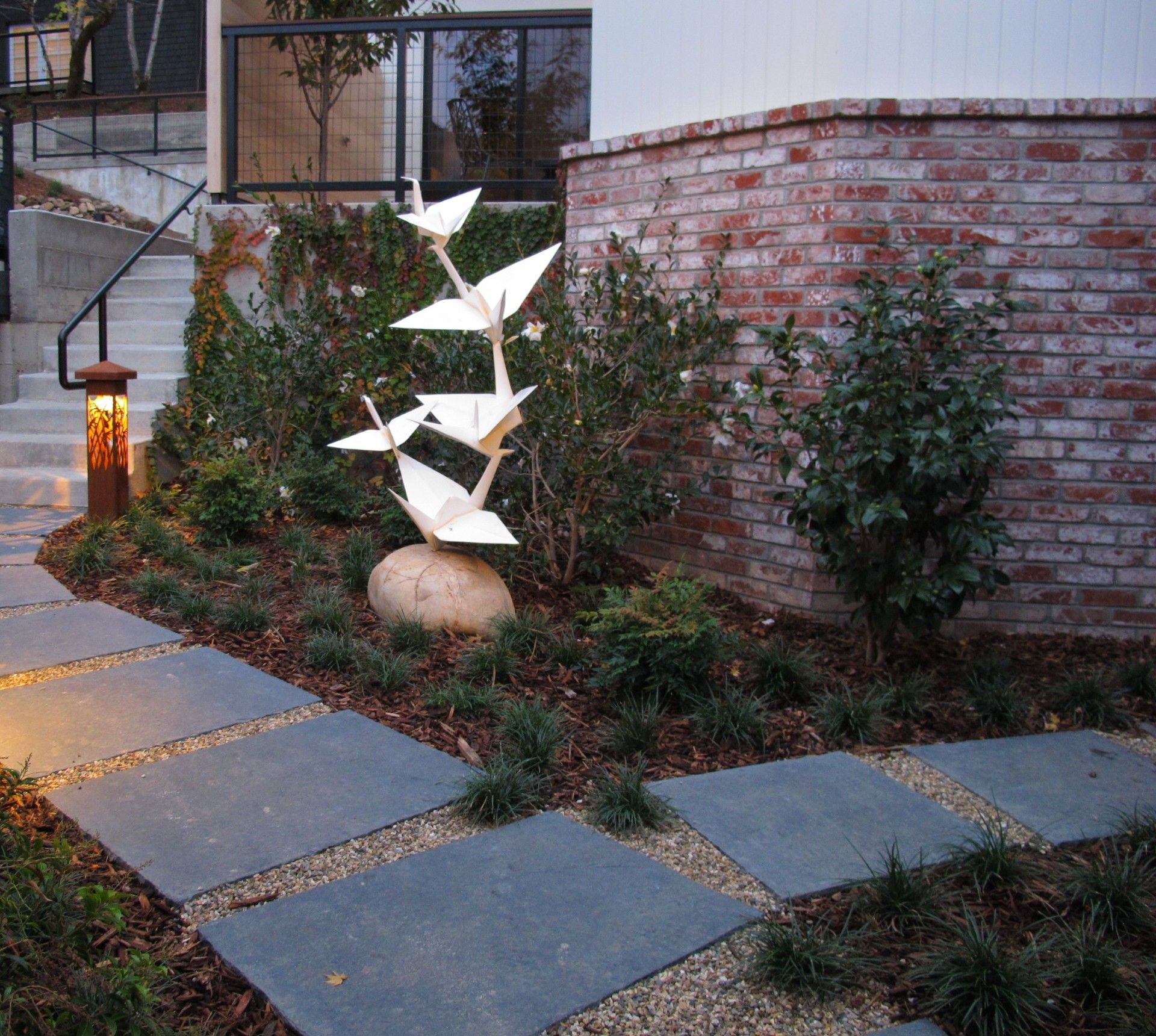 Modern garden path with stone pavers, brick wall, shrubs, lantern, and white abstract sculpture