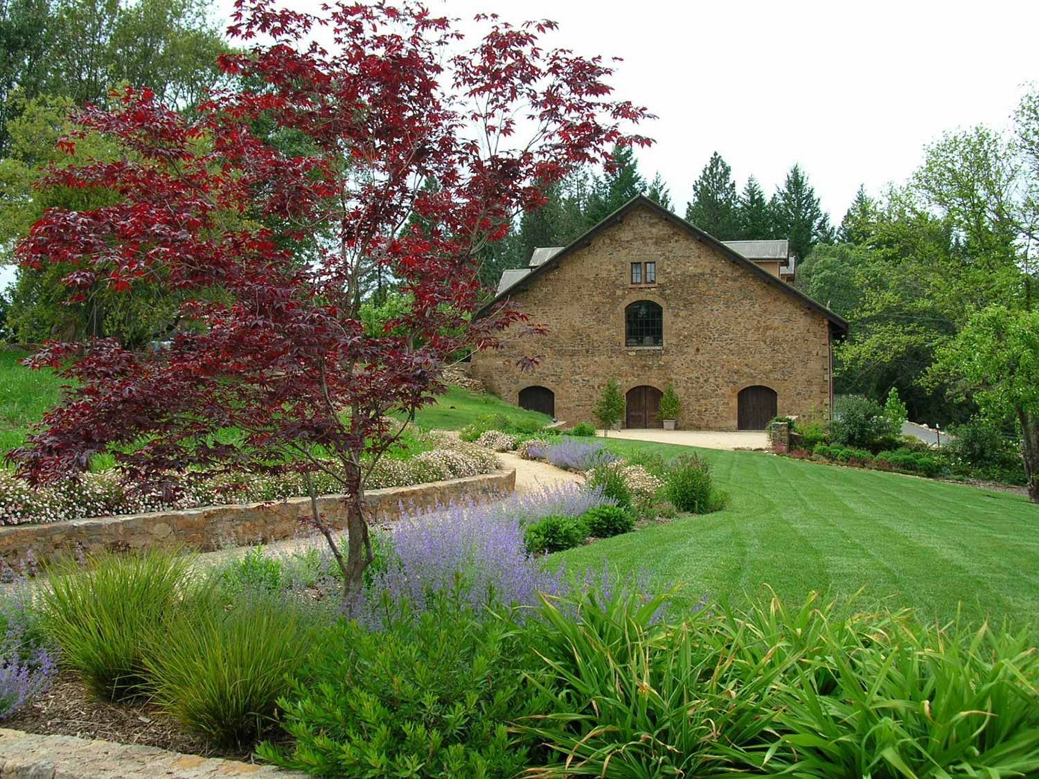 Country house with tan stone walls, red-leaf tree, and landscaped garden under a bright sky
