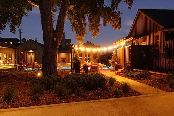 Warm outdoor courtyard at night with string lights, a large tree, and illuminated cabins along a walkway