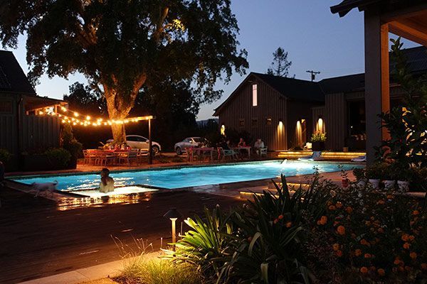 Lit resort pool at dusk with lounge chairs, glowing lights, and a dark cabin in the background