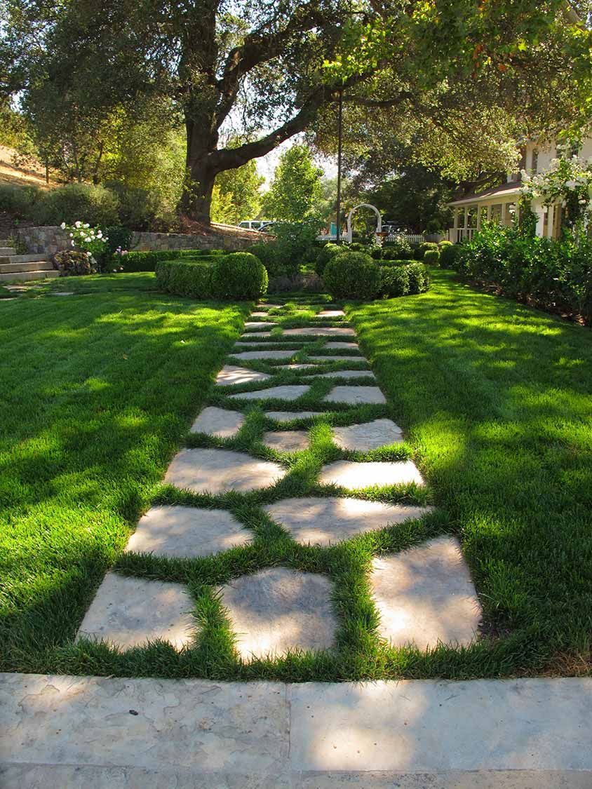 Stone path through a green garden lawn under large trees, with sunlight and flowers along the sides